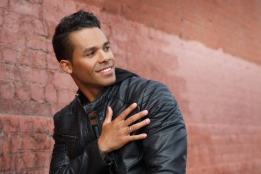 Siyah deri ceketli ve gülümseyen genç, mutlu, Latin bir adam kalbini kırmızı tuğla bir duvarın arka planına dayadı..