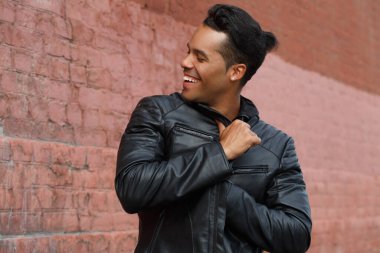 Siyah deri ceketli genç, mutlu Latin adam kırmızı tuğla bir duvarın arkasında gülümsüyor..