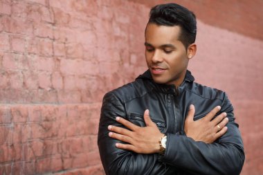 Siyah deri ceketli genç, mutlu Latin adam kırmızı tuğla bir duvarın arkasında gülümsüyor..