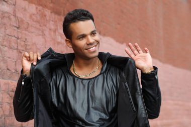 Siyah deri ceketli genç, mutlu Latin adam kırmızı tuğla bir duvarın arkasında gülümsüyor..