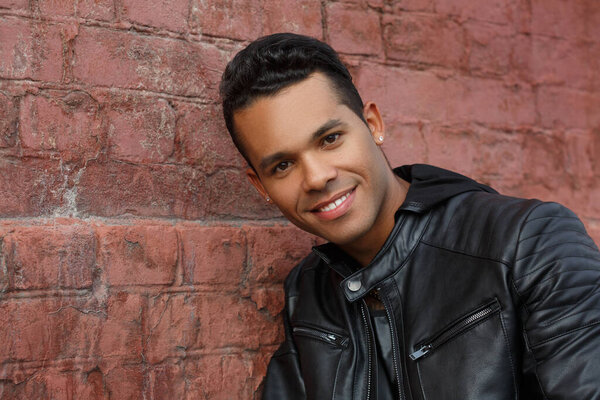 Young happy cute Latinos guy in a black leather jacket smiles against a background of a red brick wall.