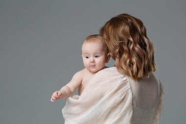Anne kollarında sevimli bir bebek tutuyor, omuzlarının üzerinden bakıyor, gri bir arka planda izole edilmiş..