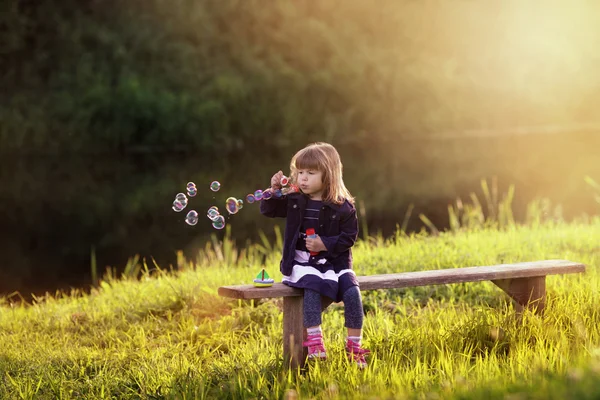 Little girl sitting on a wooden bench blows bubbles in the rays — Stock Photo, Image