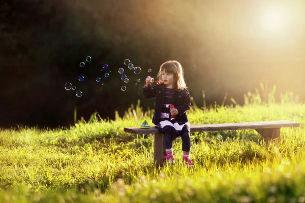 Little girl sitting on a wooden bench blows bubbles in the rays — Stock Photo, Image