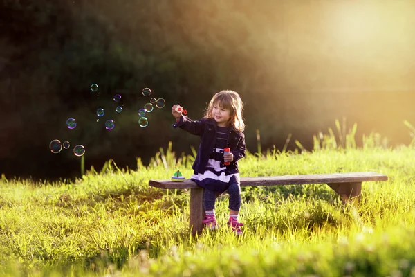 Little girl sitting on a wooden bench blows bubbles in the rays — Stock Photo, Image