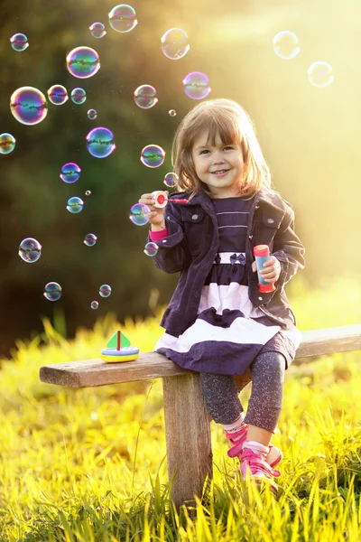 Portret van een meisje, zittend op een houten bankje blaast bubbels in th — Stockfoto