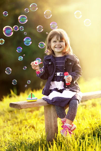 Portrét dívky sedící na dřevěné lavici fouká bublinky v th — Stock fotografie