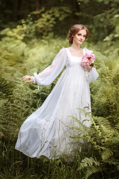 Fille sur le bord de la forêt dans une longue robe blanche volant avec un bouquet de pivoines dans les mains — Photo