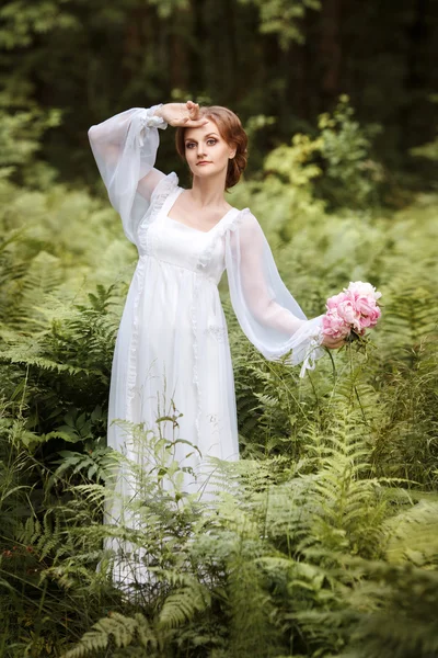 Дівчина на краю лісу в довгій білій сукні з букетом півоній в руках — стокове фото
