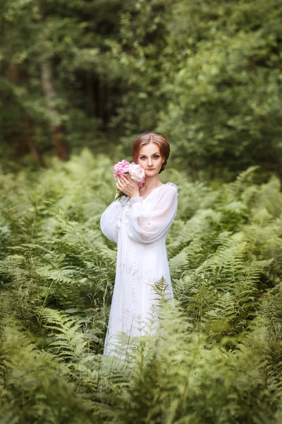 Chica en el borde del bosque en un vestido blanco largo con un ramo de peonías en las manos — Foto de Stock