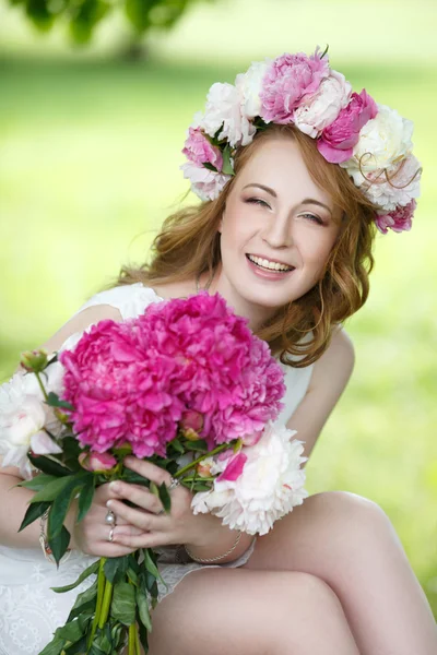 Menina feliz em uma grinalda e com um buquê de peônias na natureza — Fotografia de Stock