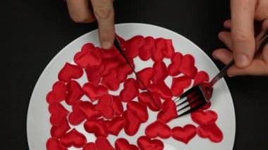 Çatal ve bıçakla kırmızı kalp yiyen biri. Kalp katili, kalp kırıcı, kaşıkçı kavramı. Siyah arka planda beyaz tabakta bir sürü kalp var.