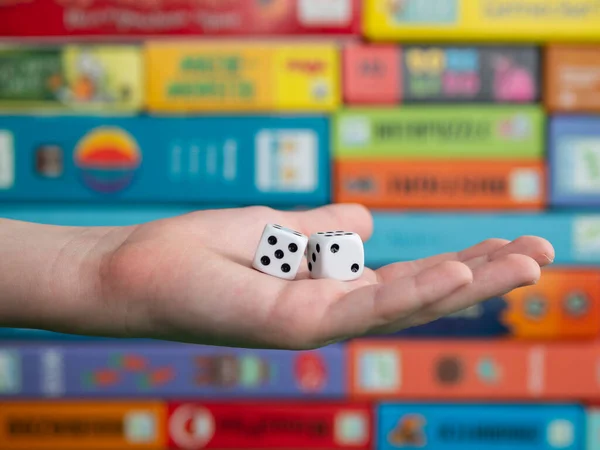 Hand with white dice on the blurred background of colorful board game boxes. Concept of entertainment during pandemic of covid-19