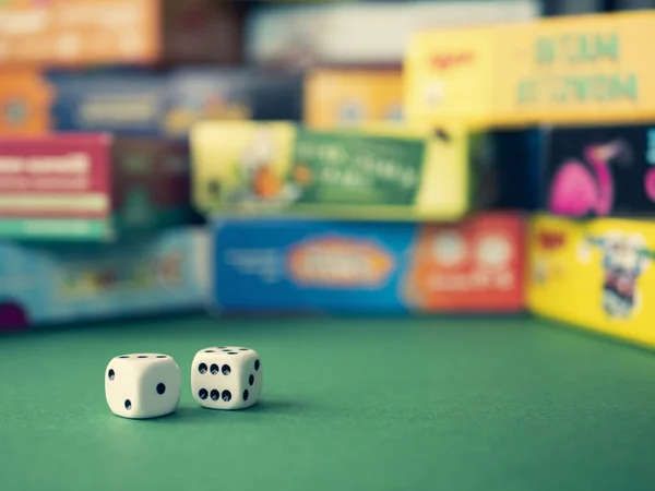 White dice on the green surface on the blurred background of colorful board game boxes. Concept of entertainment under lockdown