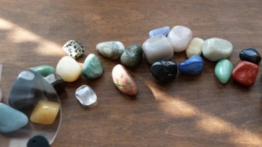 Multicolored semi-precious stones on a wooden table are being explored with a magnifying glass. Gemstones and a loupe