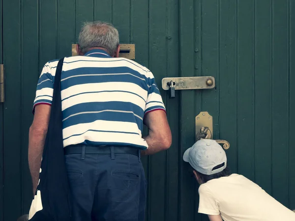 Senior and child are looking through the mail slot and keyhole, view from the back. Grandfather and grandson see something interesting. Concept of two generations, peeping and peeking