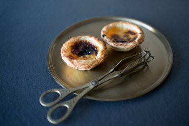 Two delicious Belem cakes (Pasteis de Belem) on a bronze tray, with serving tongs.  Portuguese sweet making tradition. Gastronomy.