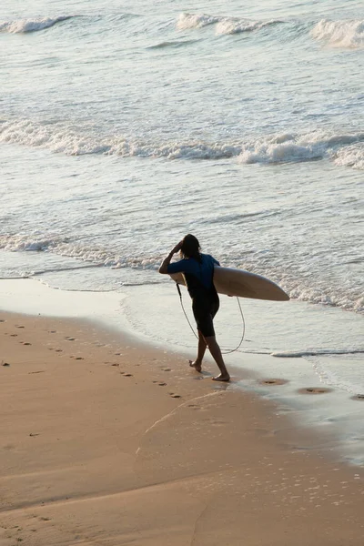 Genç adam sörf yaptıktan sonra sahilde yürüyor. Sırtında sörf tahtasıyla görülüyor. Gijon, Asturias, İspanya, Avrupa