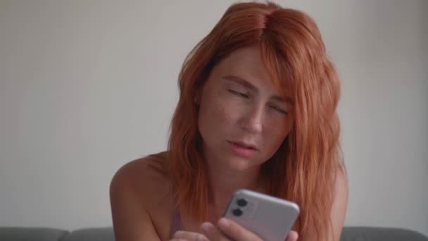 赤い髪の女の子はソファに座って、彼女の手で電話を使用します. — ストック動画