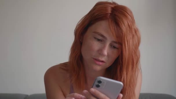 赤い髪の女の子はソファに座って、彼女の手で電話を使用します. — ストック動画