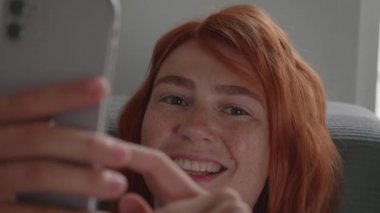 Kızıl saçlı bir kız telefonu elleriyle kullanıyor, kanepede yatıyor..