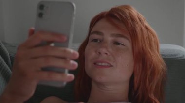 Kızıl saçlı bir kız telefonu elleriyle kullanıyor, kanepede yatıyor..