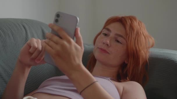 A girl with red hair uses the phone with her hands, lying on the couch. — Stock Video