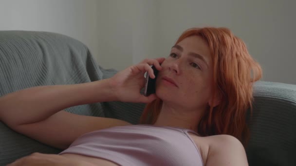 赤い髪の女の子はソファの上に横たわって、彼女の手で電話を使用します. — ストック動画