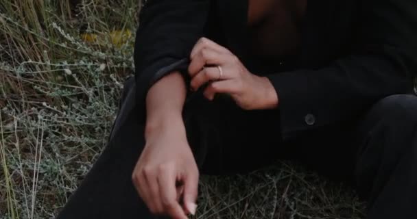 Una chica sexy negra en una chaqueta está sentada en la hierba por la noche en la playa. — Vídeos de Stock