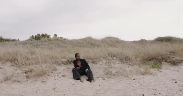 Una chica sexy negra con chaqueta está sentada en la playa. Amanecer. — Vídeos de Stock