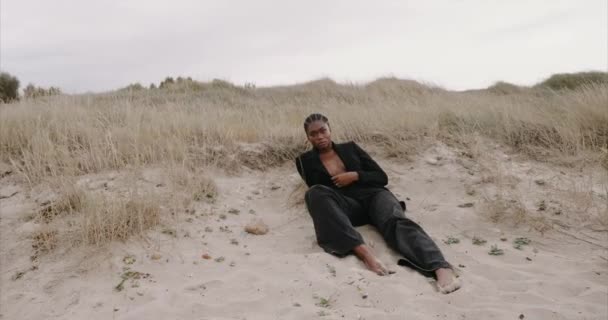 Een zwart sexy meisje in een jasje zit op het strand. Zonsopgang. — Stockvideo