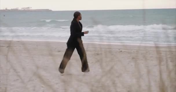 Una chica sexy negra con chaqueta corre a lo largo de la playa. Amanecer. — Vídeos de Stock