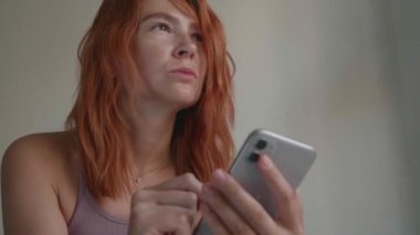 Kızıl saçlı bir kız telefonu elleriyle kullanıyor, kızgın bir şekilde koltukta oturuyor.