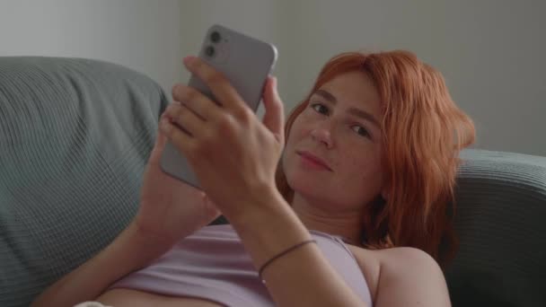 Una chica de pelo rojo usa el teléfono con las manos, acostada en el sofá. — Vídeos de Stock