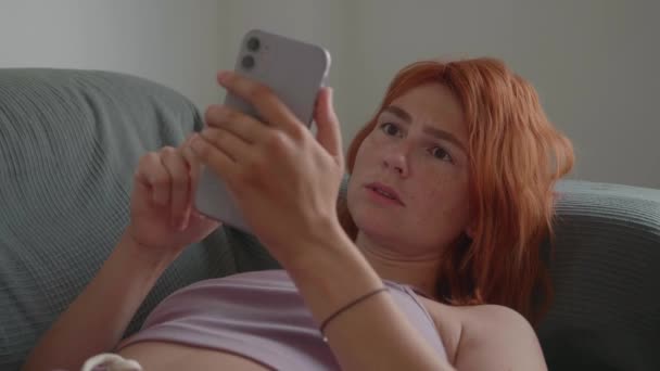 Ένα κορίτσι με κόκκινα μαλλιά χρησιμοποιεί το τηλέφωνο με τα χέρια της στον καναπέ.. — Αρχείο Βίντεο