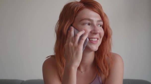 Seorang gadis dengan rambut merah menggunakan telepon dengan tangannya, duduk di sofa. — Stok Video