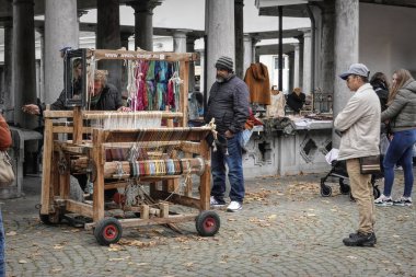 Bruges, Flanders, Belçika 10 18 2025 Kadın açık hava pazarında ahşap dokuma tezgahında çalışıyor. Alışverişçiler, piyasanın eşsiz atmosferiyle çevrili olarak yakınlarda geziniyorlar..