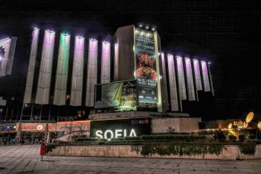 Sofya 'daki aydınlık binalar gece gökyüzüne karşı parıldıyor. Sahne, 'Sofya' tabelasıyla parlayan mimari güzelliği yakalıyor, canlı bir şehir manzarası yaratıyor, yaya ile tamamlanmış..