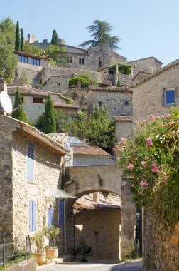Düşük açılı bir açıdan çekilen fotoğrafta bir yamaçta yuva yapmış taştan bir köy görülüyor. Binalar, bazı mavi panjurlar ve canlı çiçeklerle sıkıca gruplandırılmış. Kemer yolları gölgeli şeritlere çıkıyor.. 