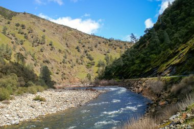 Dağlık bir vadide, kayalık kıyıları ve yemyeşil bitkileri olan bir nehir kıvrılır. Açık mavi gökyüzü, doğanın dinginliğinin özünü yakalayan sakin sahneyi güçlendirir..