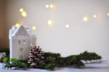 Yeşil ladin dallarının yanında pencereleri olan beyaz seramik evler ve çiçek ışıklarıyla bulanık bir zemin üzerinde kar kaplı koniler, yumuşak odaklı yatay görüntü ve metin için yer