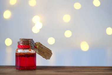 Üzerinde kırmızı-altın sıvısı olan küçük bir cam kavanoz ve bir altın etiket, tahtadan bir masanın bulanık bir masasında duruyor. Üzerinde bir çelenk, yatay bir görüntü, yumuşak bir odak ve metin için bir yer var.