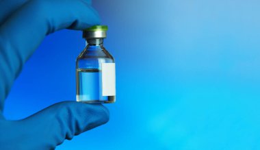 Beyaz etiketli ve içinde şeffaf sıvı olan ampuller, mavi tıbbi eldivendeki el bir aşı ile bir balon tutar, yumuşak odaklı yatay bir görüntü, geniş bir afiş, metin için bir yer