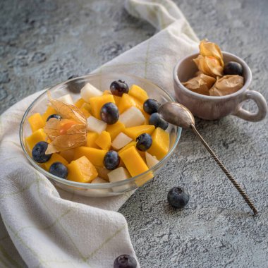 Mango parçalı meyve salatası ve cam kasede yaban mersini. Beyaz bir bez üzerinde Phisalis. Gri arkaplanda bir kopya alanı olan meyve salatası .