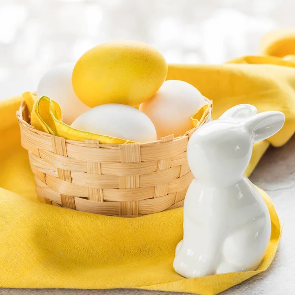 Colored Easter egg and white eggs in a basket and rabbit on a yellow cloth with a copy space. Yellow Easter egg colored by turmeric powder .Natural dye for eggs. Easter eggs painted with natural dyes