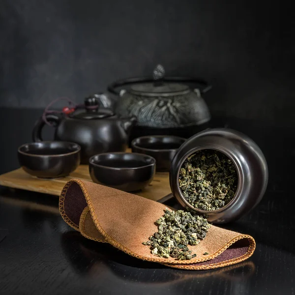 Green tea brew in a jar , black teapot , cups on a wooden black  table .  Green tea brew on a brown  cloth.   Tea ceremony on a black background .