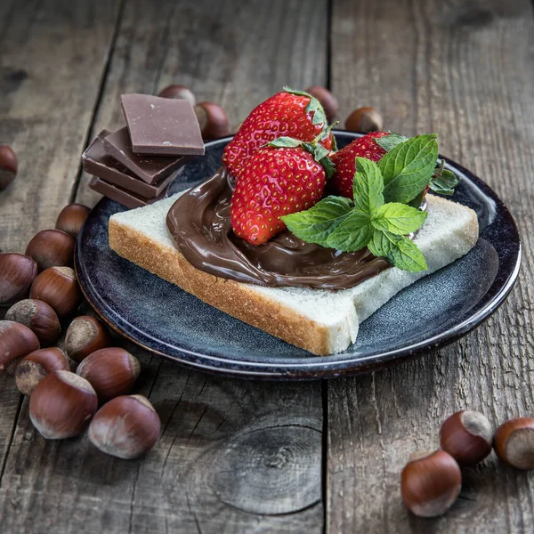 A piece of bread with chocolate and nuts paste on a blue plate . Heazelnuts, strawberry , mint leaves , bread with  chocolate paste and chocolate pieces  on a wooden background .