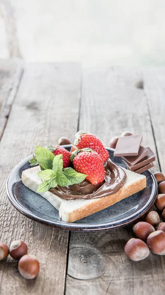 A piece of bread with chocolate and nuts paste on a blue plate with a copy space  . Heazelnuts, strawberry , mint leaves , bread with  chocolate paste and chocolate pieces  on a wooden background .