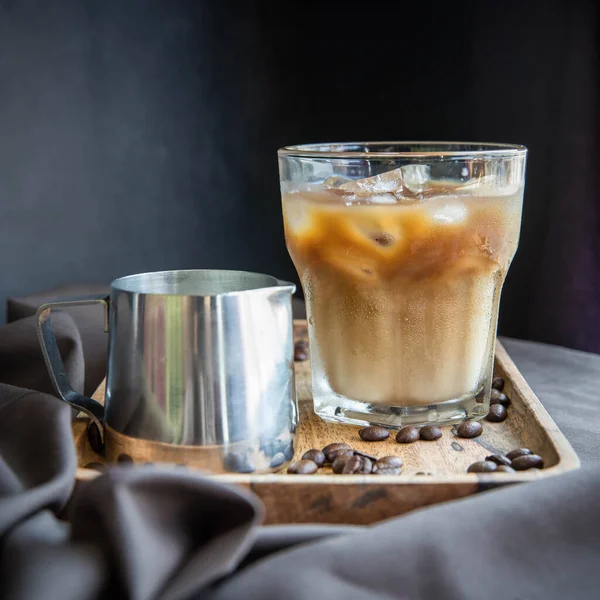 An iced coffee with milk and a milk pitcher  on a wooden tray and black background .  Refreshning . Roasted coffee beans .