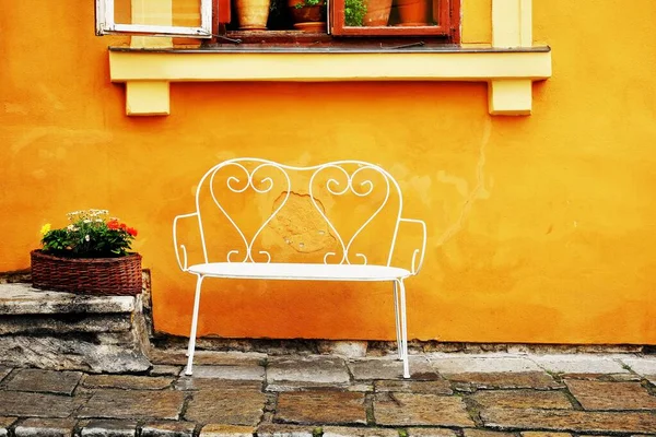 Street photo. White bench under house window. There are flowers in the window and on the street. The yellow color of the house is dominant. Street still life photo after rain.
